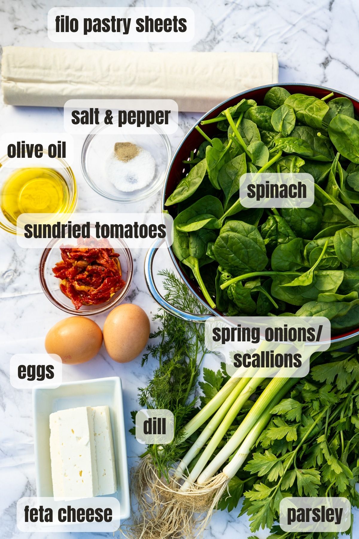 All the ingredients needed to make a Greek spinach and feta filo pie including filo pastry, feta cheese, eggs, fresh herbs, olive oil, sundried tomatoes and seasoning.