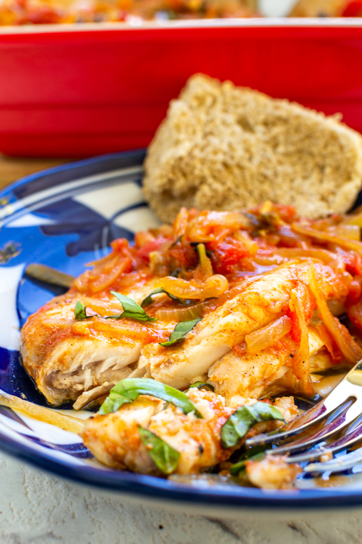 A closeup of Greek white fish with tomatoes and onions on a blue patterned dish with fresh basil and a fork.