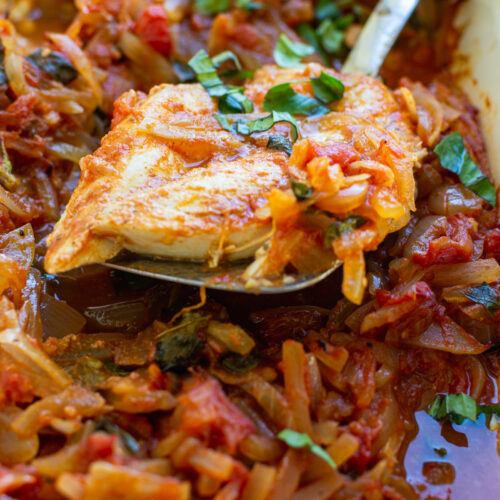 Closeup of someone spooning Greek fish baked in tomatoes and onions from a red baking dish.