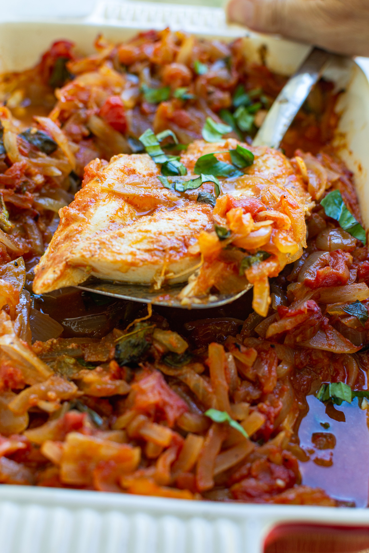 Closeup of someone spooning Greek fish baked in tomatoes and onions from a red baking dish.