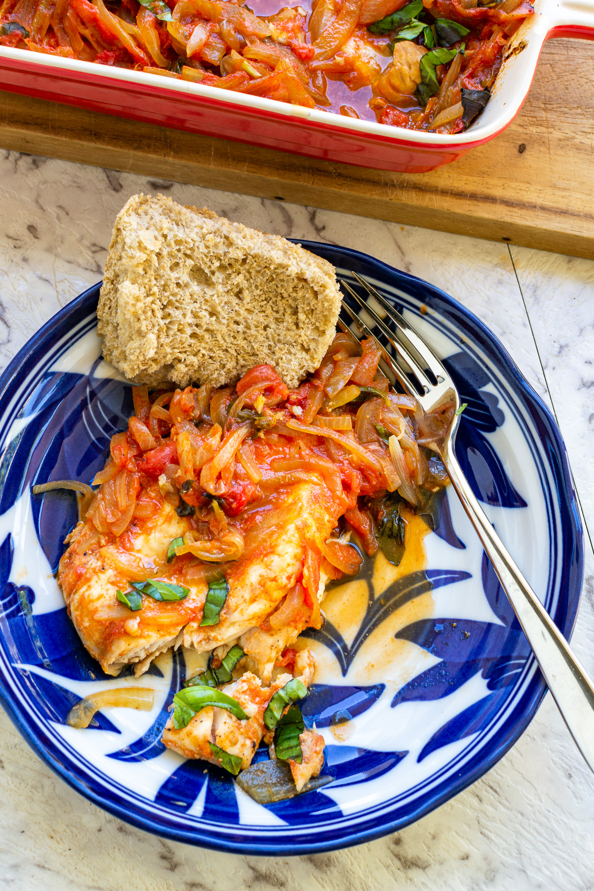 Overhead view of a piece of Greek baked fish with tomatoes and onions and basil on a patterned blue and white plate with a fork and a piece of bread with red baking dish behind.