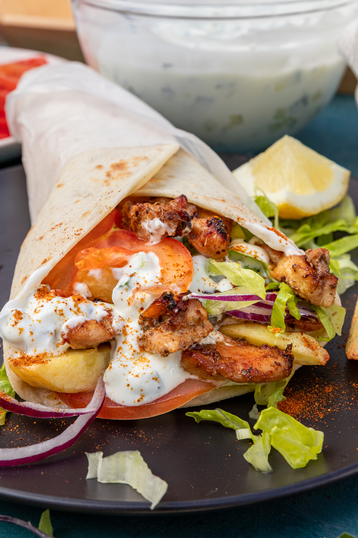 closeup of the inside of a Greek chicken gryo wrap with chicken thigh, tzatziki, onion, tomatoes and lettuce on a black plate with a bowl of tzatziki in the background.