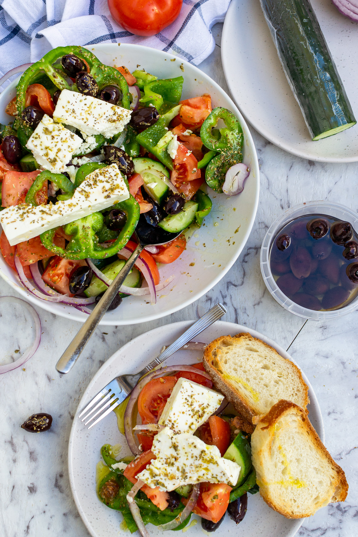 A portion of Greek salad on a plate and the rest of the salad in a bowl from above on a marble background.