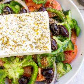 Part of a big Greek salad in a white bowl with a big slice of feta cheese and oregano on top from above.