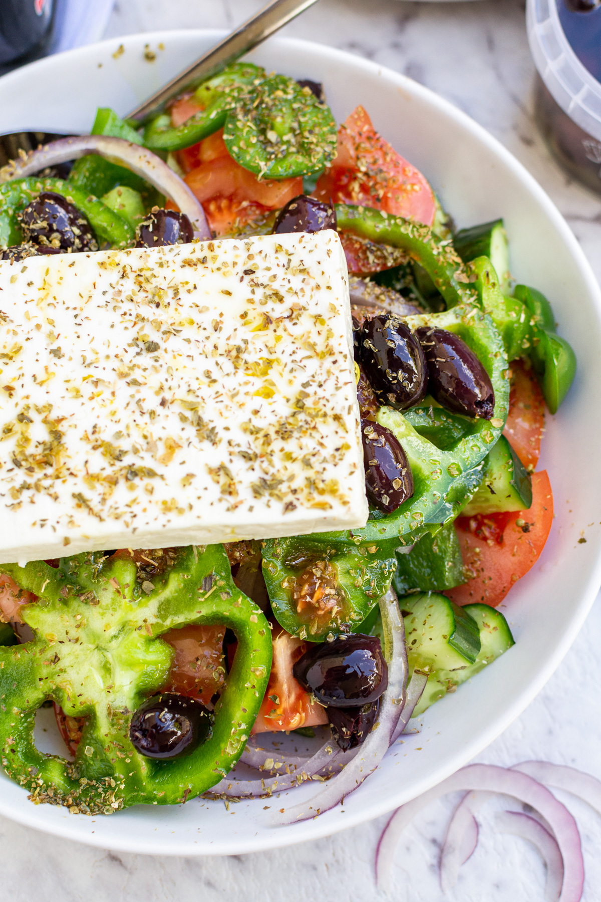 Part of a big Greek salad in a white bowl with a big slice of feta cheese and oregano on top from above.