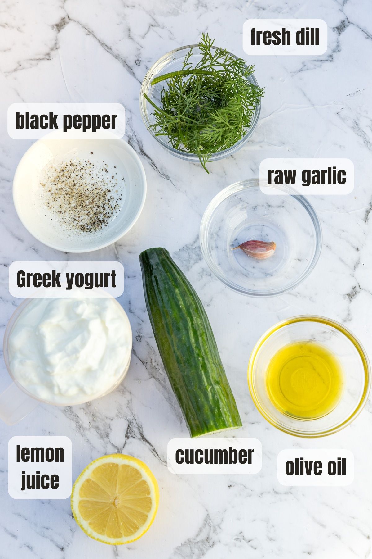 All the ingredients for tzatziki labelled on a marble background including Greek yogurt, lemon juice, cucumber, olive oil, black pepper, fresh dill and a garlic clove.