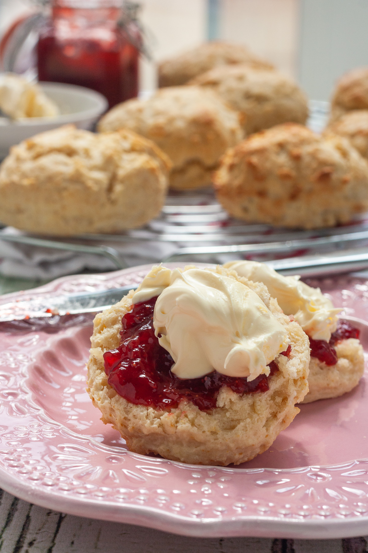 A closeup of a lemonade scone topped with jam and cream on a decorative pink plate with more scones, jam and cream in the background.