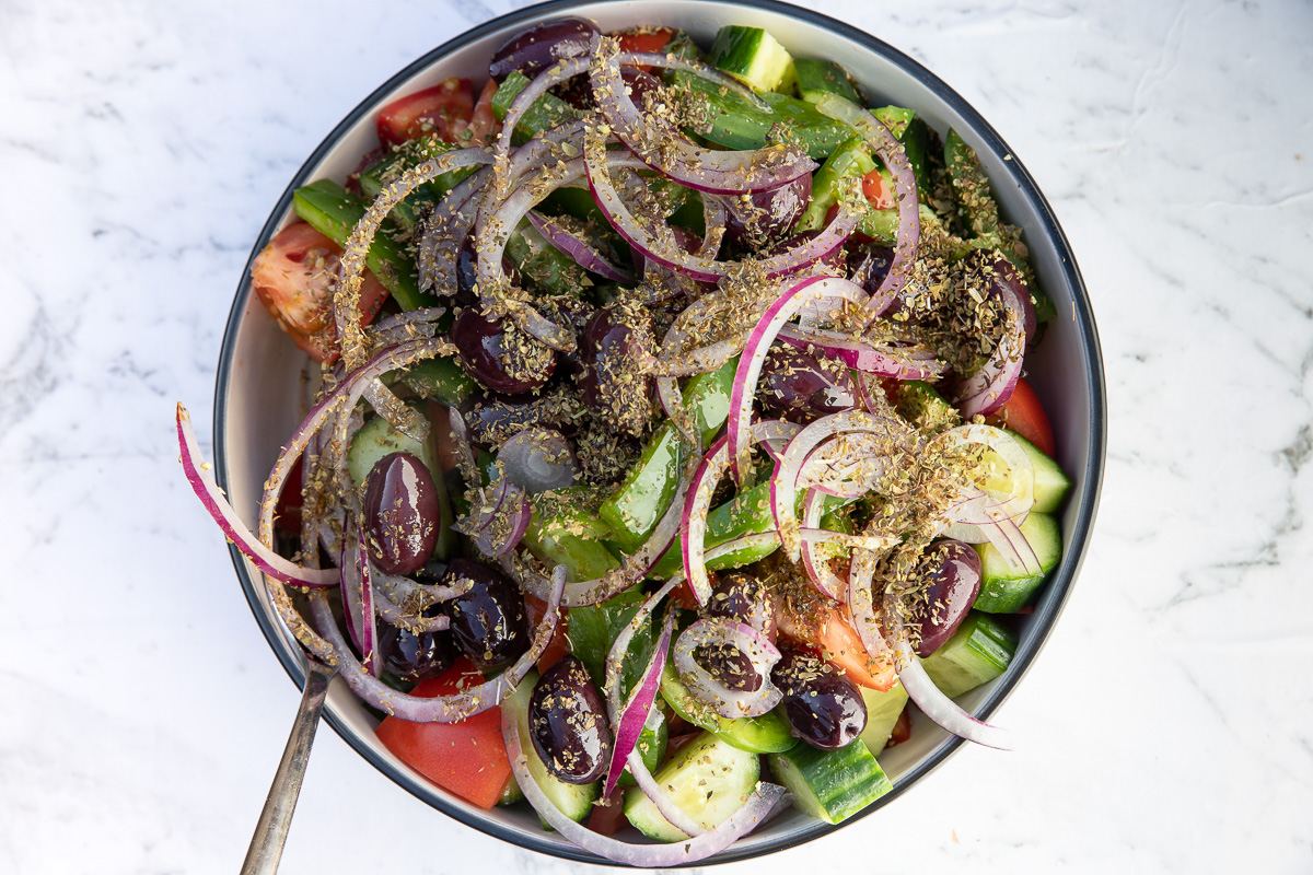 All ingredients for Greek salad apart from the feta in a big bowl with a spoon and on a marble background.