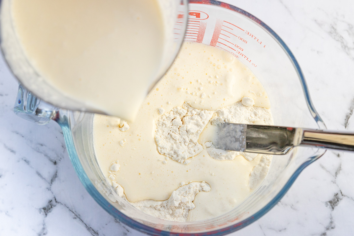 Overhead view of someone pouring liquid into flour in a glass jug and with a dinner knife for stirring on a marble background.