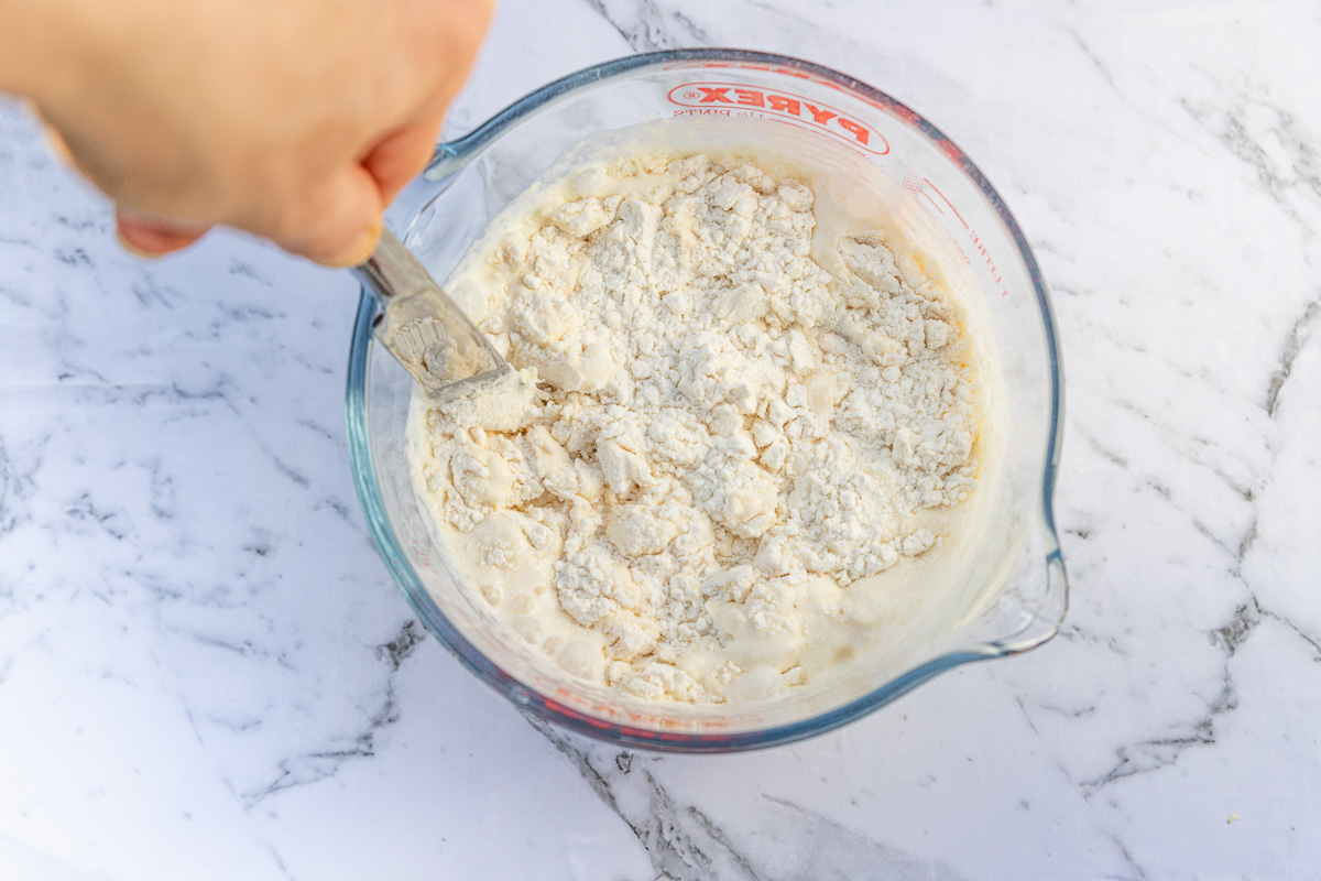 Someone stirring lemonade and cream into flour to make lemonade scones in a Pyrex jug on a marble background.