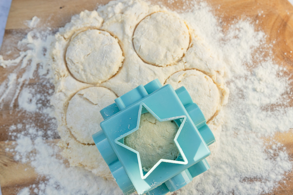 Overhead view of cutting out round scones with a round blue pastry cutter on a wooden board with flour.