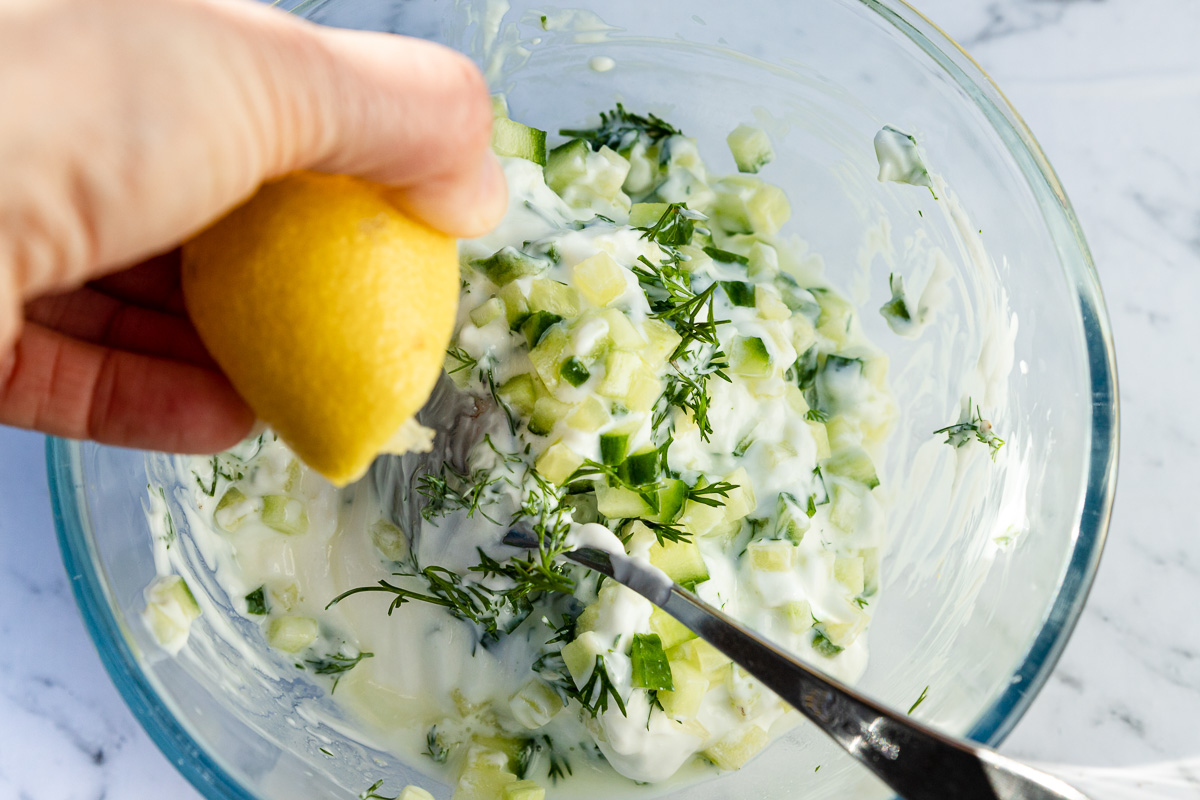 closeup of someone squeezing half a lemon into a bowl of all the ingredients for tzatziki in a glass bowl with a spoon from above.