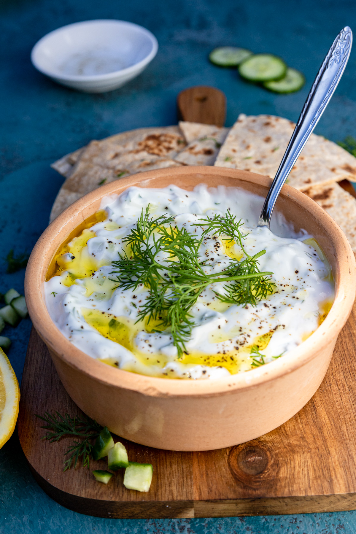 A brown ceramic dish of Greek tsatziki on a wooden board and blue background with slices of pita bread behind it.