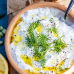 A closeup of a brown ceramic bowl of authentic Greek tsatziki with a spoon and with fresh dill and olive oil on top and on a wooden board and blue background.