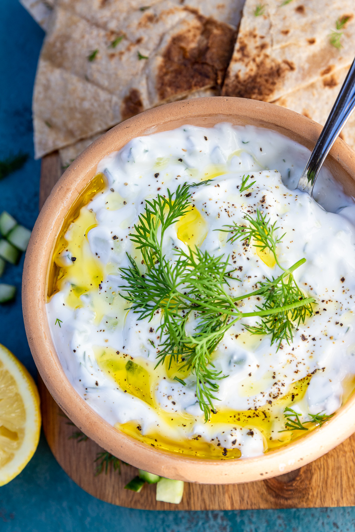 A closeup of a brown ceramic bowl of authentic Greek tsatziki with a spoon and with fresh dill and olive oil on top and on a wooden board and blue background.