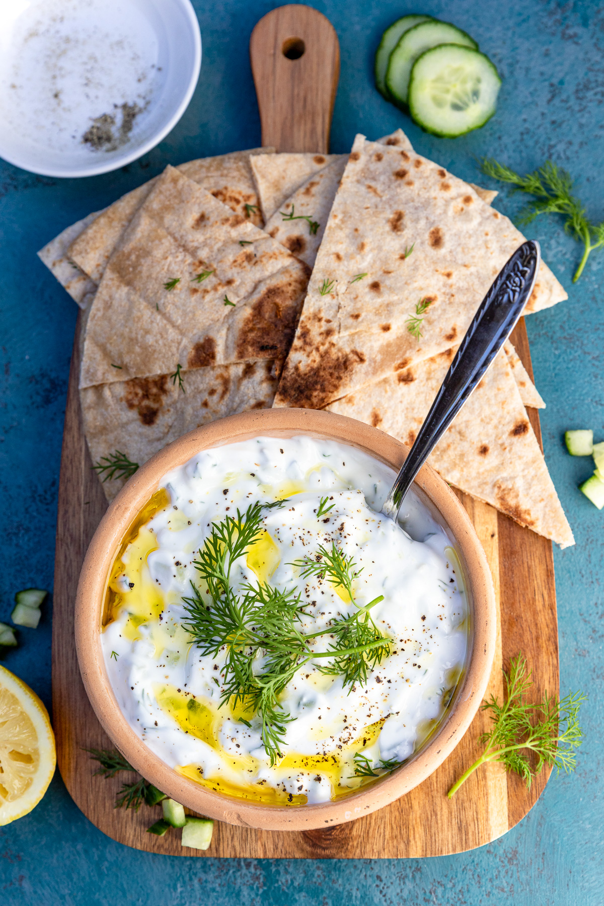 A bowl of Greek tzatziki in a ceramic dish garnished with dill, pepper and olive oil and on a wooden board with sliced pita bread.