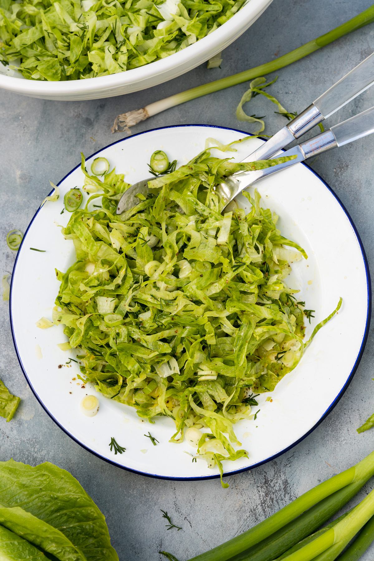 Overhead of a chopped lettuce salad and salad servers in a white dish with blue rim on a grey background with ingredients all around it.