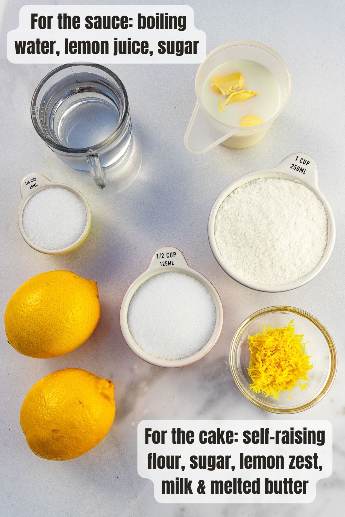 Overhead view of all the ingredients needed to make an easy lemon pudding on a marble background, including 2 lemons, lemon zest, sugar, self-raising flour, milk and butter.