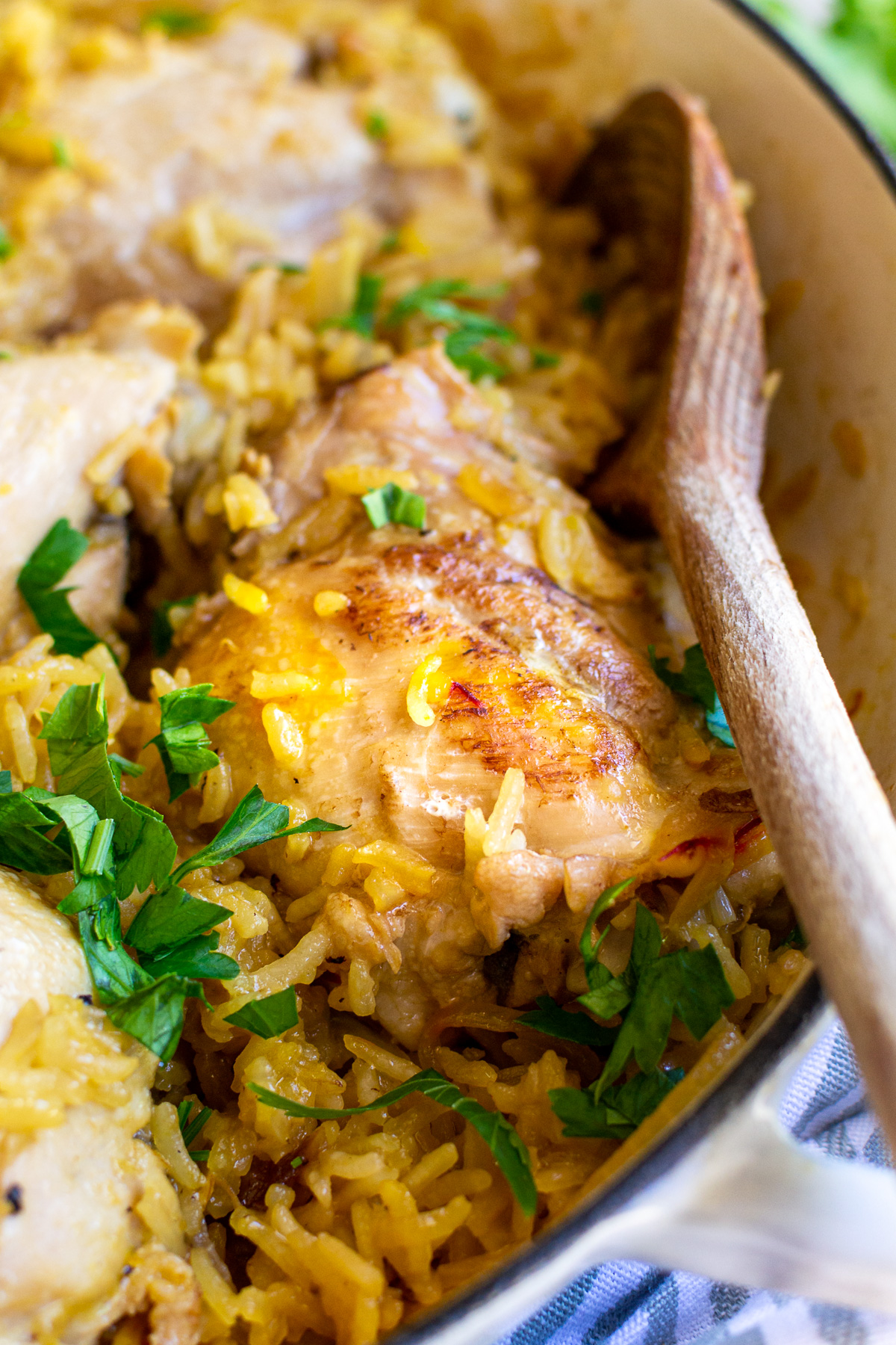 Closeup of stovetop chicken and rice in a cast iron pan on a blue striped tea towel and with a wooden spoon in it.