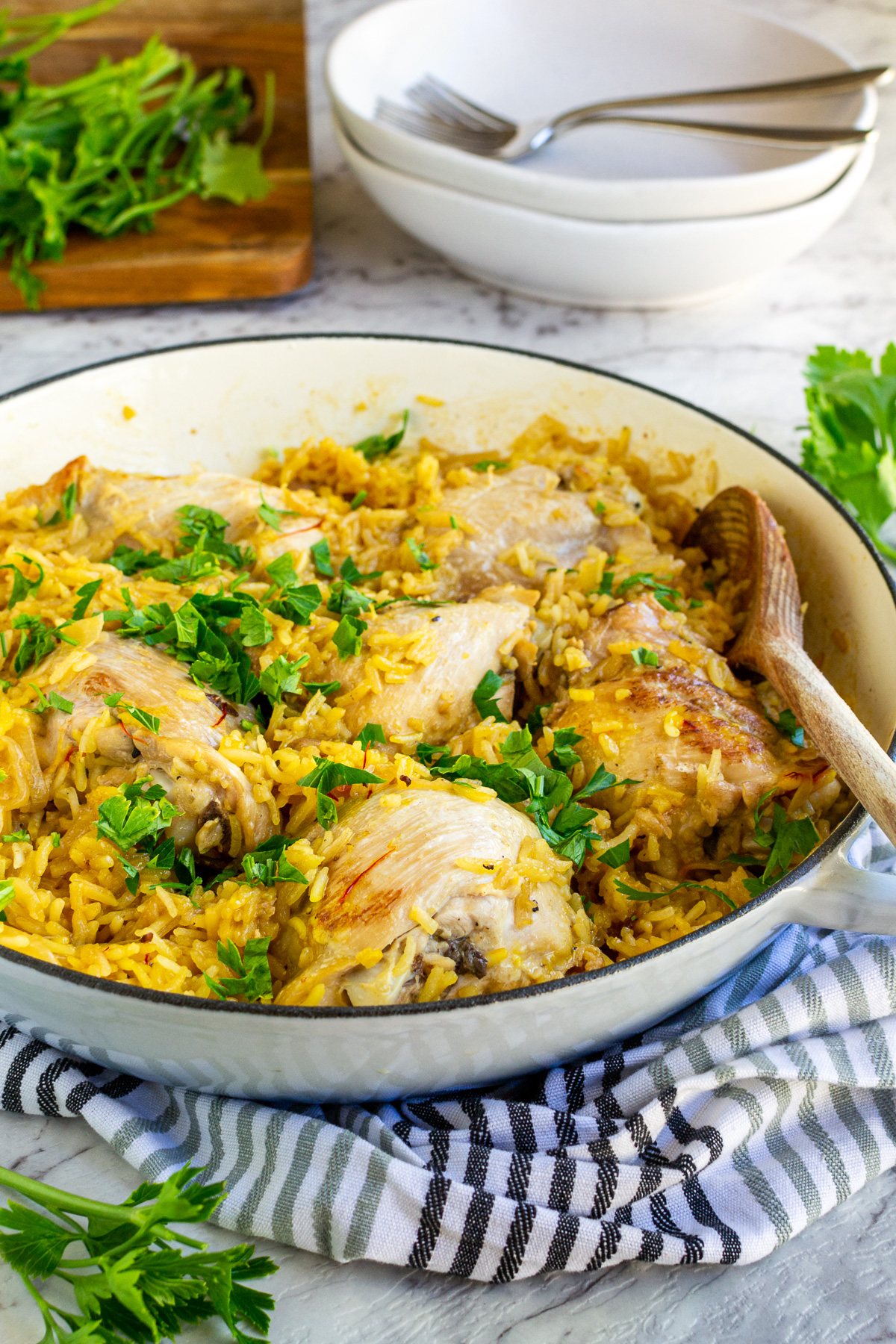 Stove top chicken and rice in a blue cast iron pan sprinkled with fresh parsley with a wooden spoon in and on a grey striped tea towel and on a marble background.