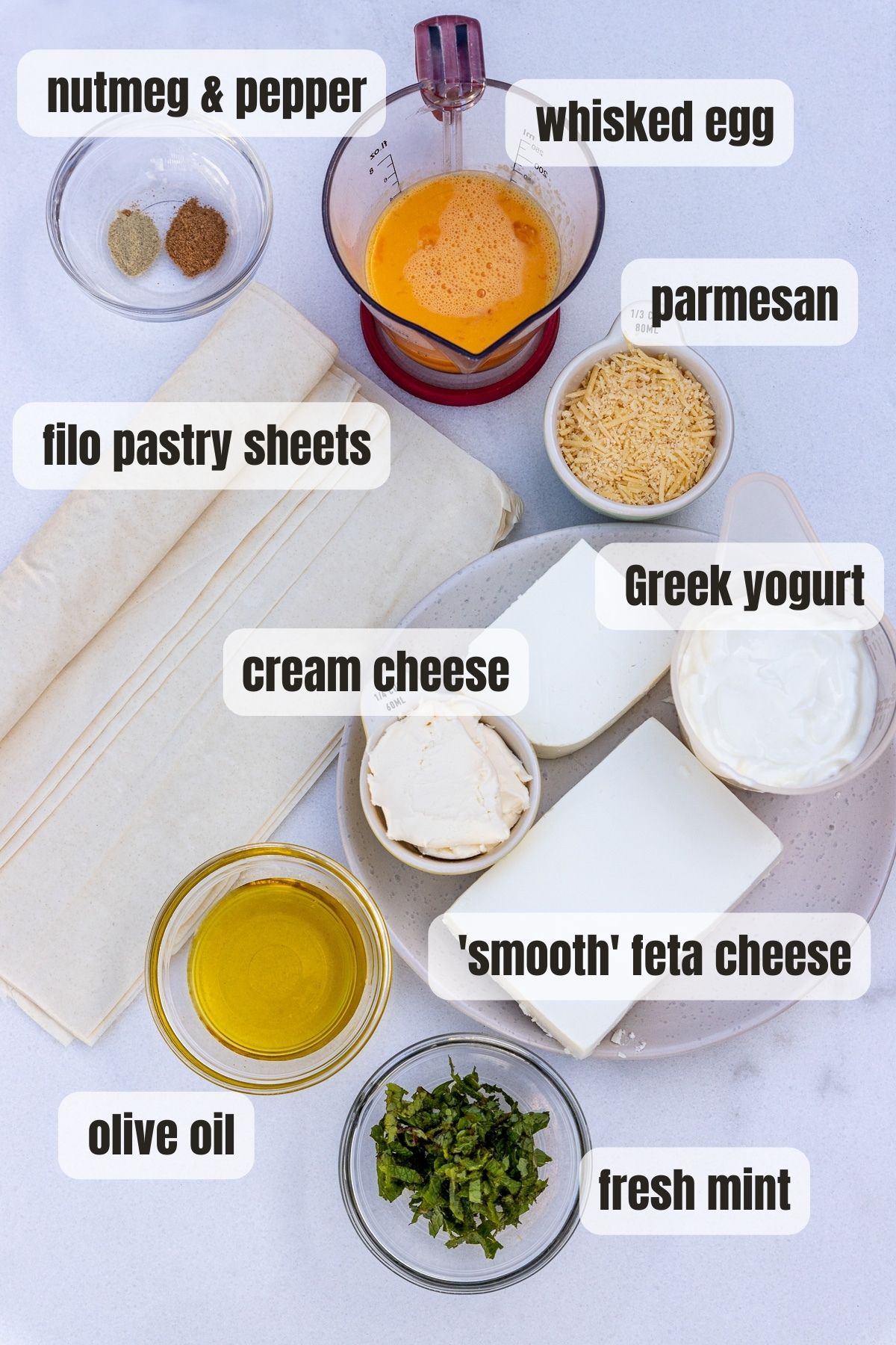 Overhead view of all the ingredients needed to make tiropitakia labelled with the following: olive oil, fresh mint, smooth feta cheese, cream cheese, Greek yogurt, whisked egg, nutmeg and pepper, parmesan and filo pastry sheets.