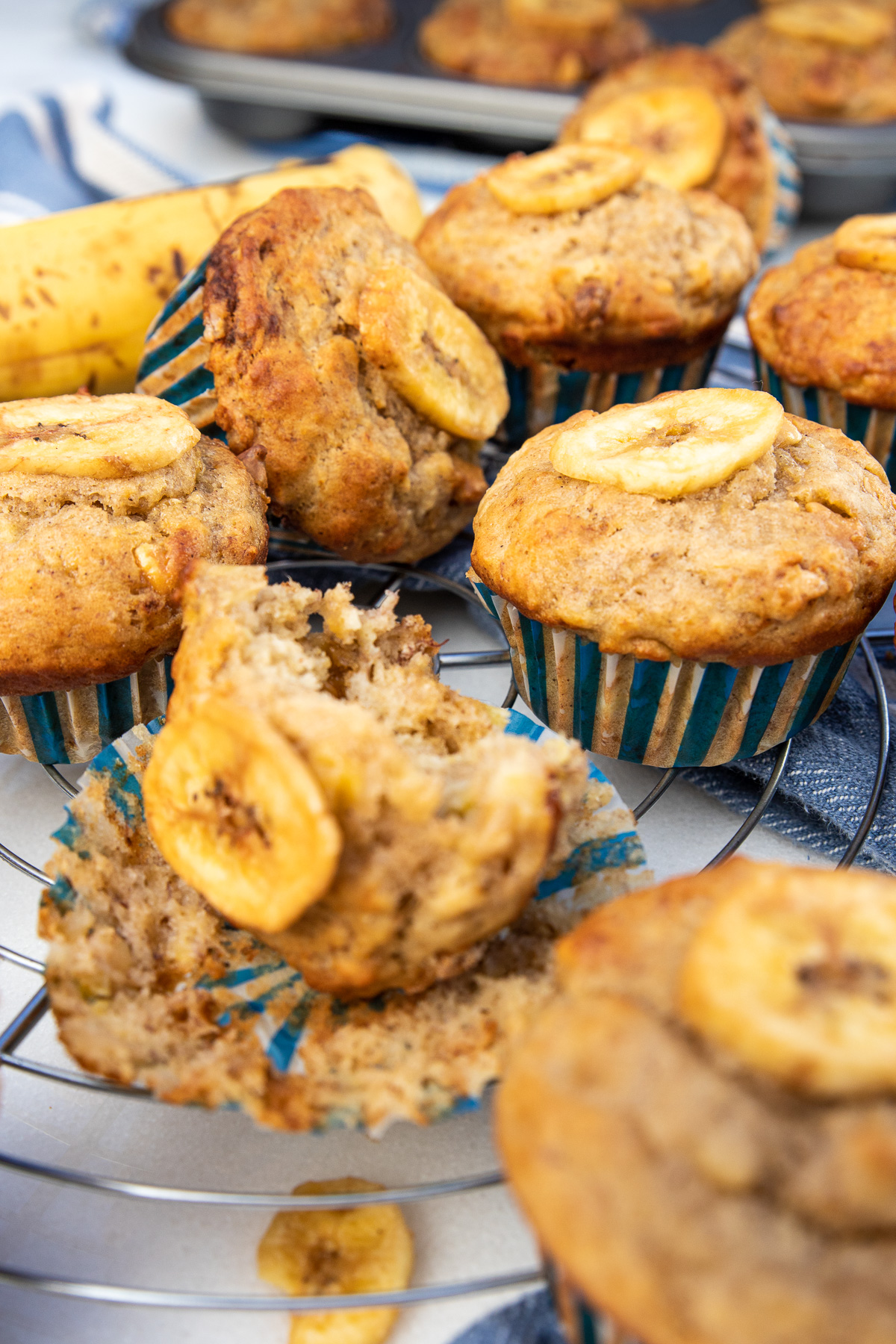 A closeup of Greek yogurt banana muffins on a cooling rack with a blue and white tea towel and ripe banana in the background.