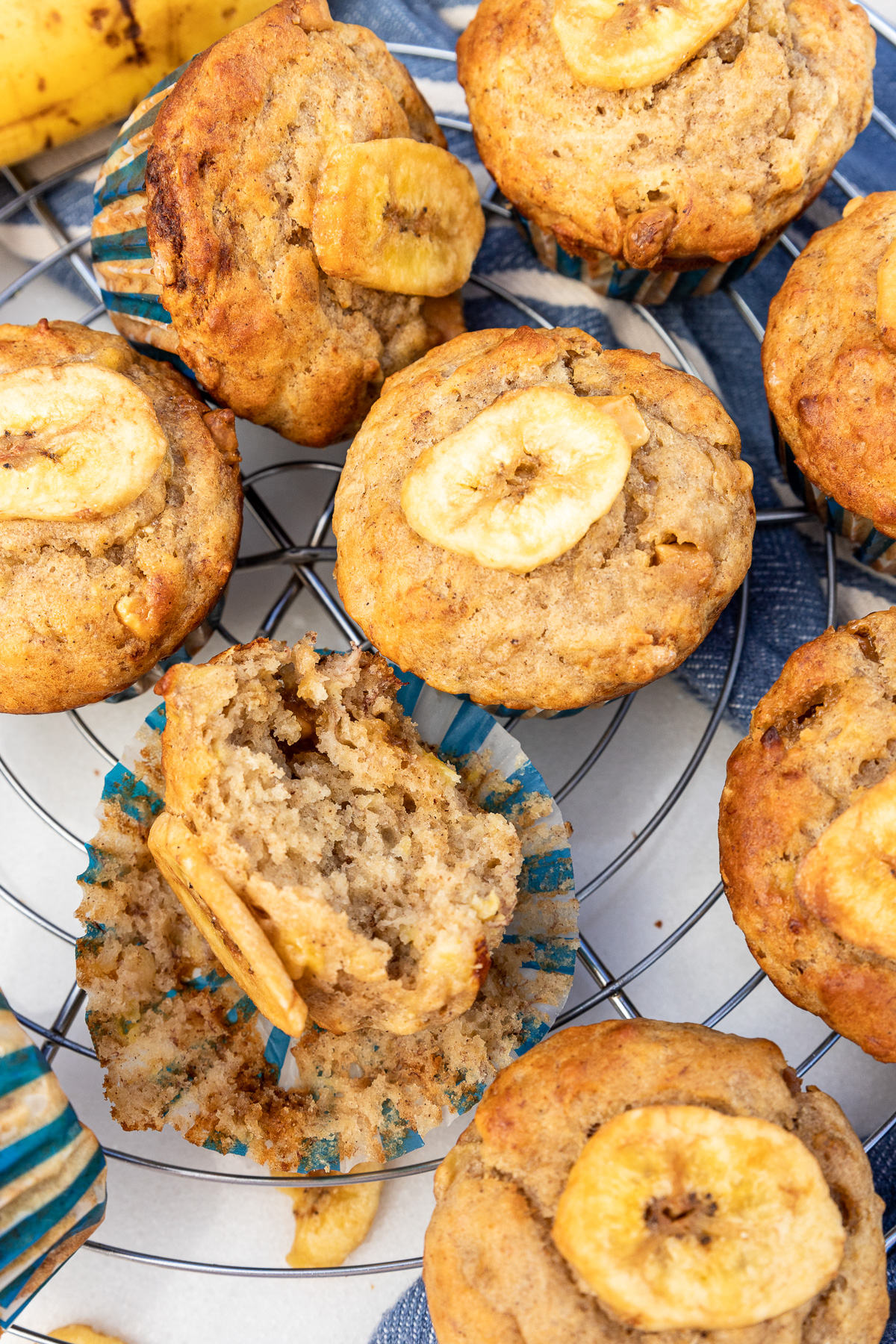 Overhead closeup of Greek yogurt banana muffins on a wire cooling rack with blue tea towel and with one muffin opened up to see the inside.