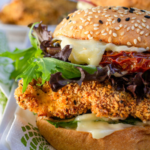 Closeup of the left side of an air fryer crispy chicken burger on a green and white napkin and rectangular white plate with more burgers and chicken in the background.