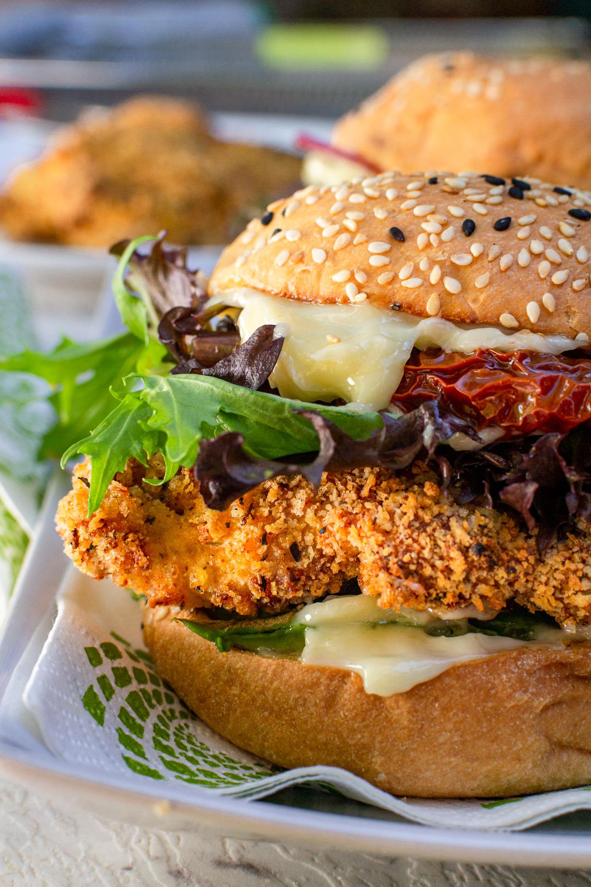 Closeup of the left side of an air fryer crispy chicken burger on a green and white napkin and rectangular white plate with more burgers and chicken in the background.