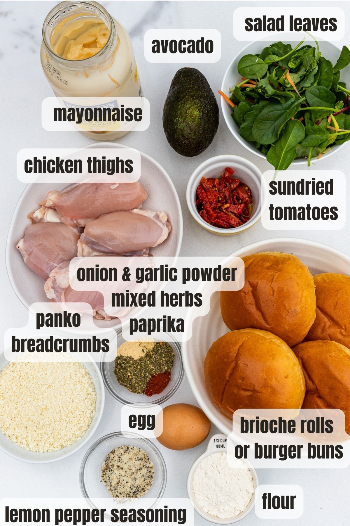 Overhead of all the ingredients to make an air fryer crispy chicken burger including lemon pepper seasoning, flour, chicken thighs, panko breadcrumbs, egg, brioche rolls, sundried tomato, mayonnaise, onion and garlic powder, mixed herbs and paprika.