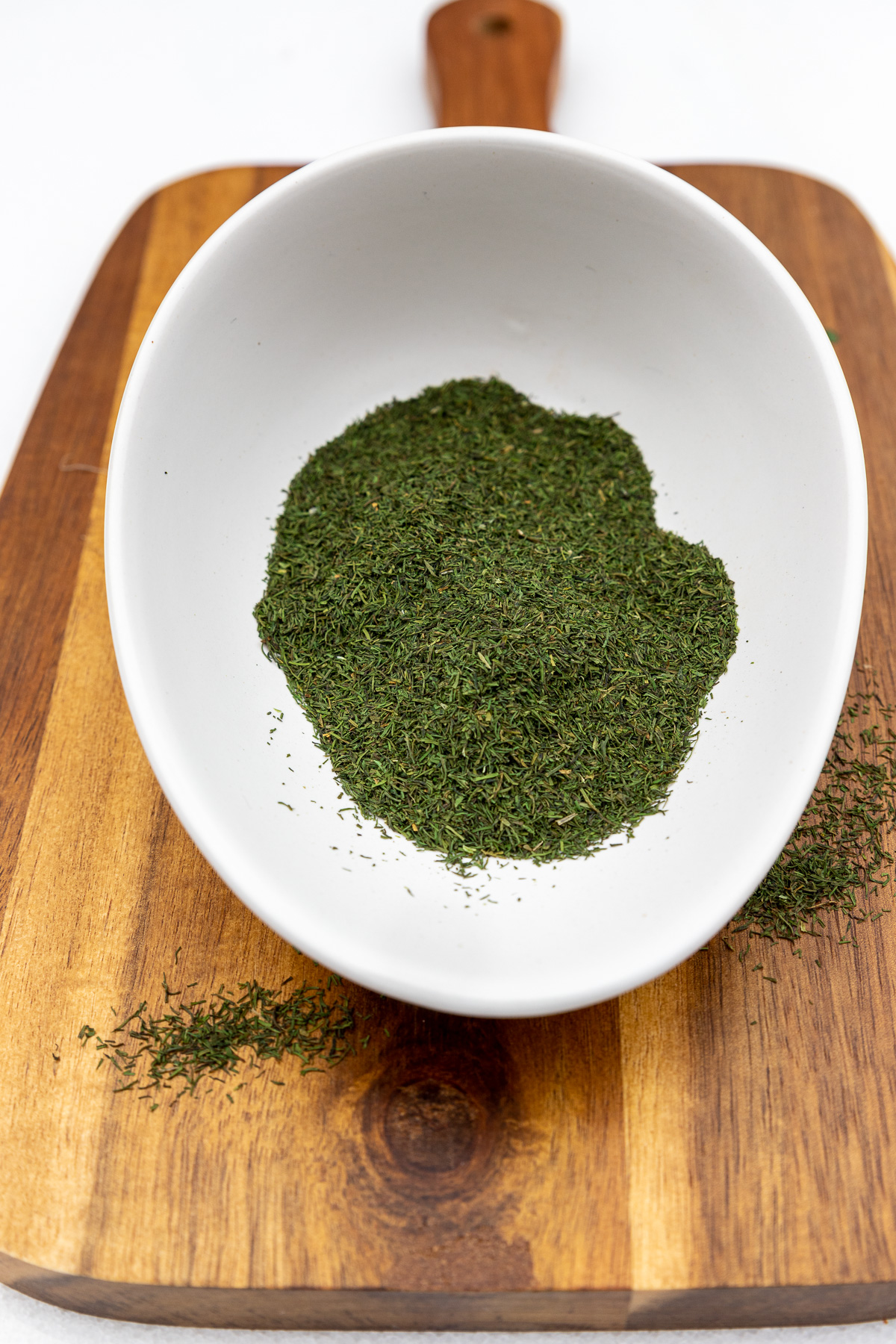 Dried dill in a small white bowl on a wooden chopping board.