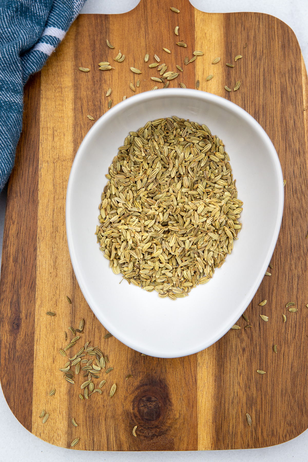 Dried fennel seeds in a small white bowl on a wooden chopping bowl and with the edge of a blue and white tea towel on the left.