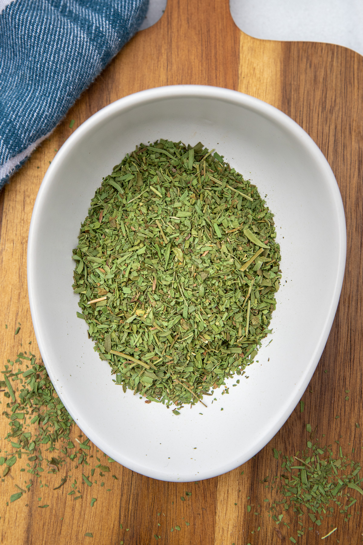 Dried tarragon in a white bowl on a wooden board next to a white and blue tea towel.