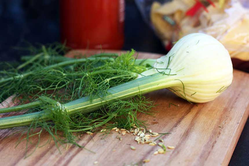 A bulb of fresh fennel complete with fronds on a wooden chopping board with other ingredients in the background.
