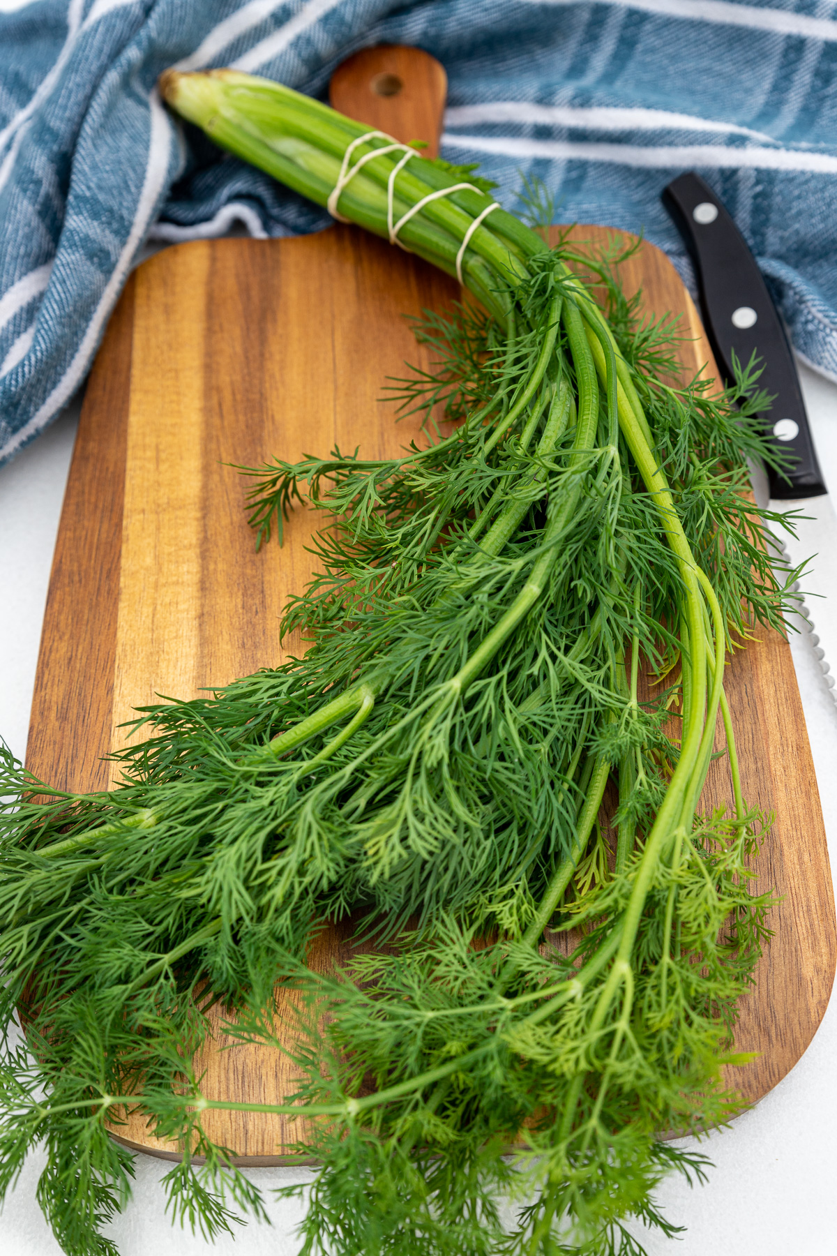 A bunch of fresh green dill on a wooden chopping board with a knife and a blue and white tea towel behind.