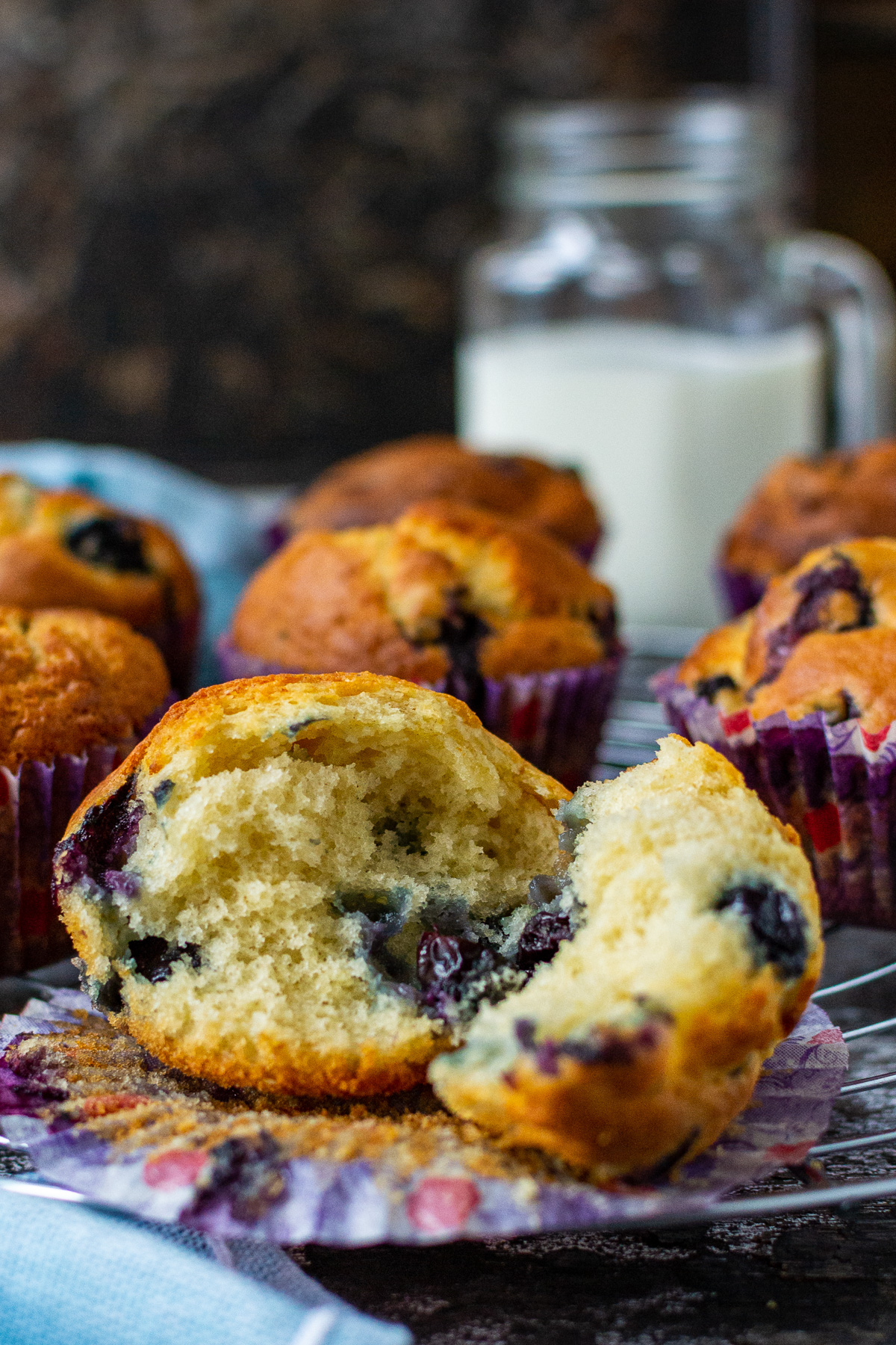 The inside of a fluffy Greek yogurt blueberry muffin on a paper liner and dark background with a light blue tea towel in front and more muffins and milk in the background.