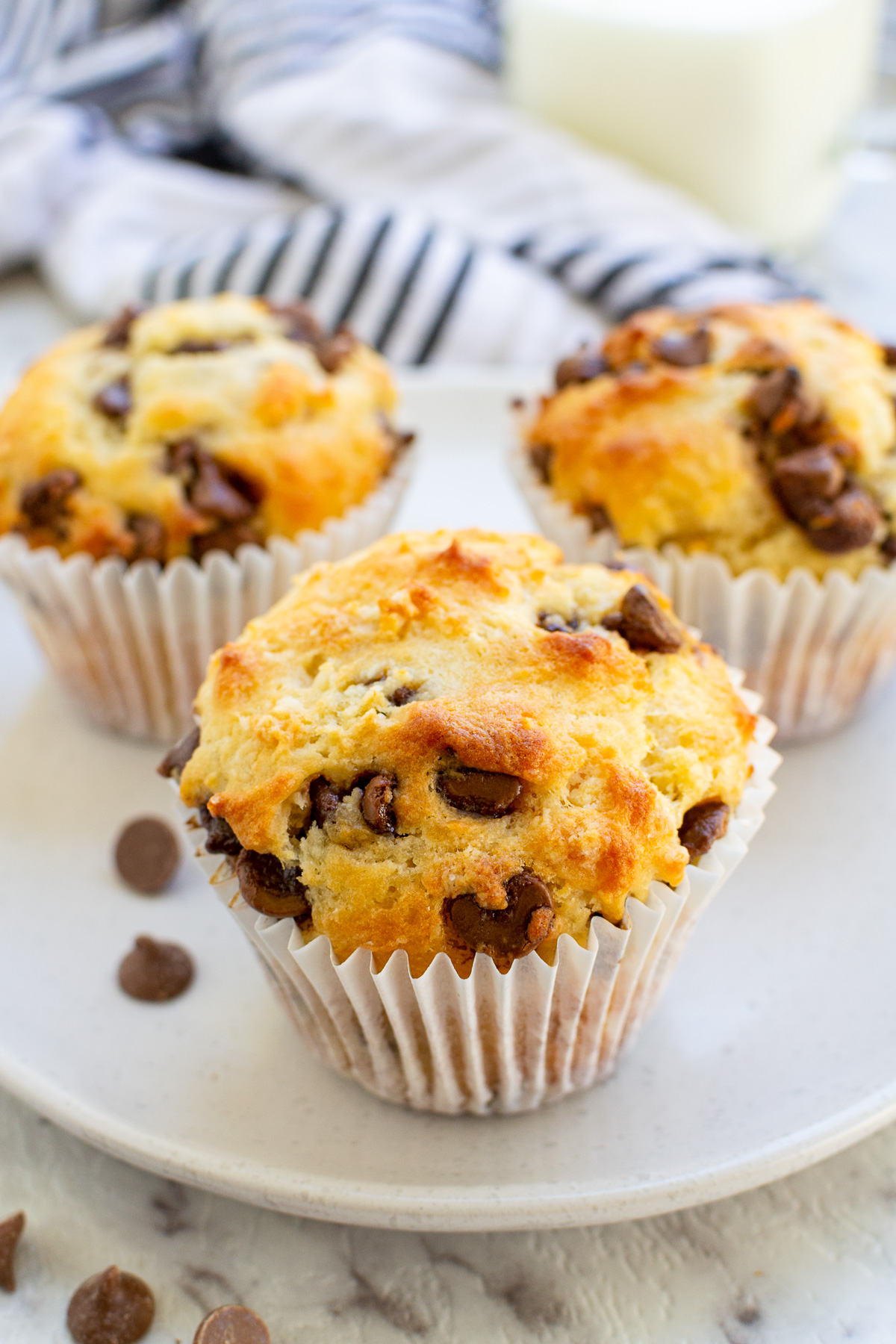 Closeup of a Greek yogurt choc chip muffin on a pale beige plate and marble surface with more cakes and a striped blue tea towel behind.