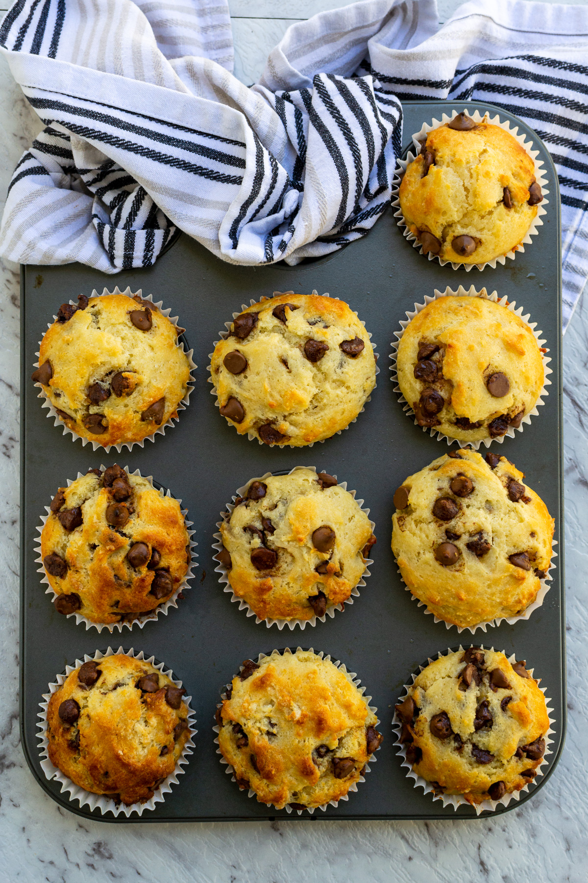 Overhead view of a pan of just baked Greek yogurt choc chip muffins on a marble background with a blue and grey striped tea towel at the top.