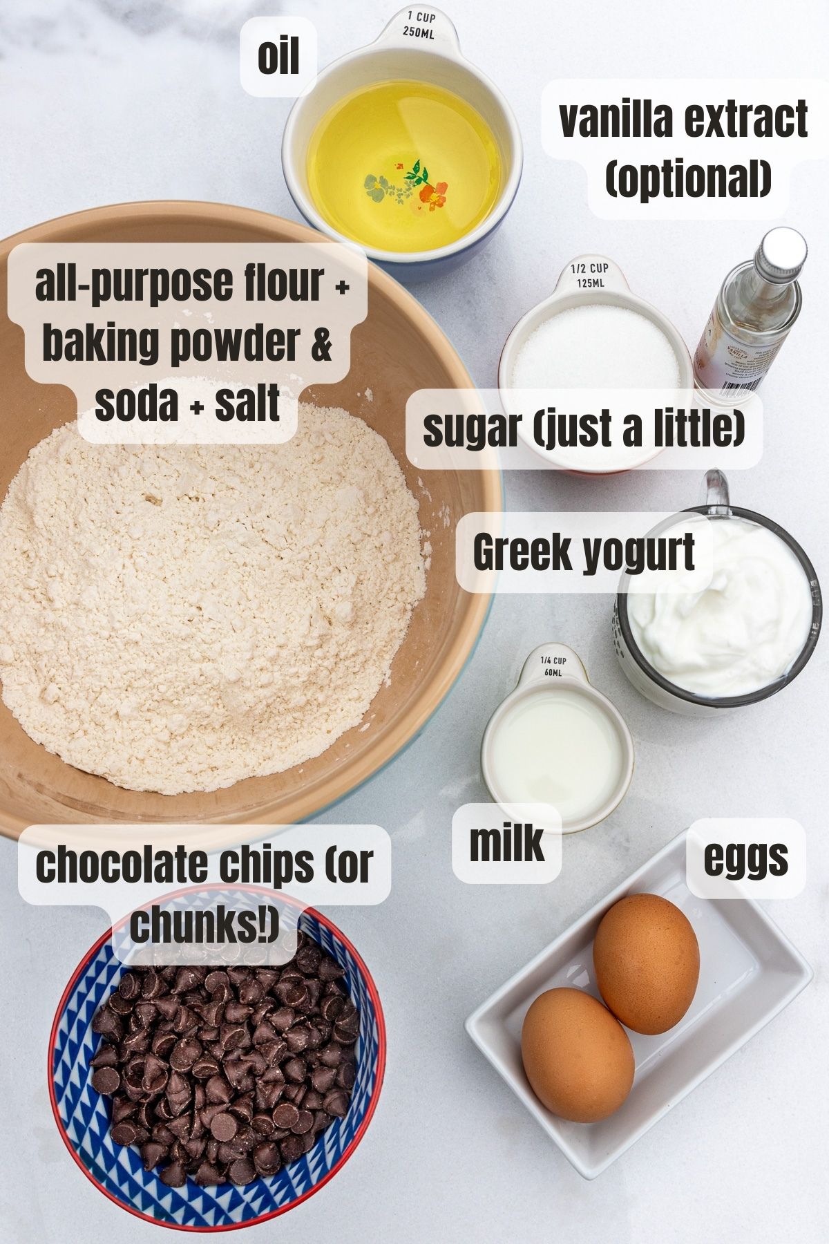 Overhead view of all the ingredients needed to make Greek yogurt chocolate chip muffins including chocolate chips, eggs, milk, Greek yogurt, flour, baking powder, baking soda and salt, vanilla extract, oil and sugar.