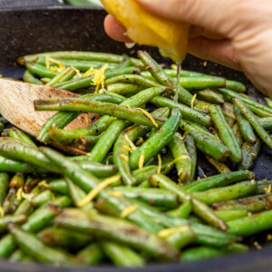 Closeup of someone squeezing half a lemon into a black cast iron pan of sauteed frozen green beans with a pale green tea towel in the background.