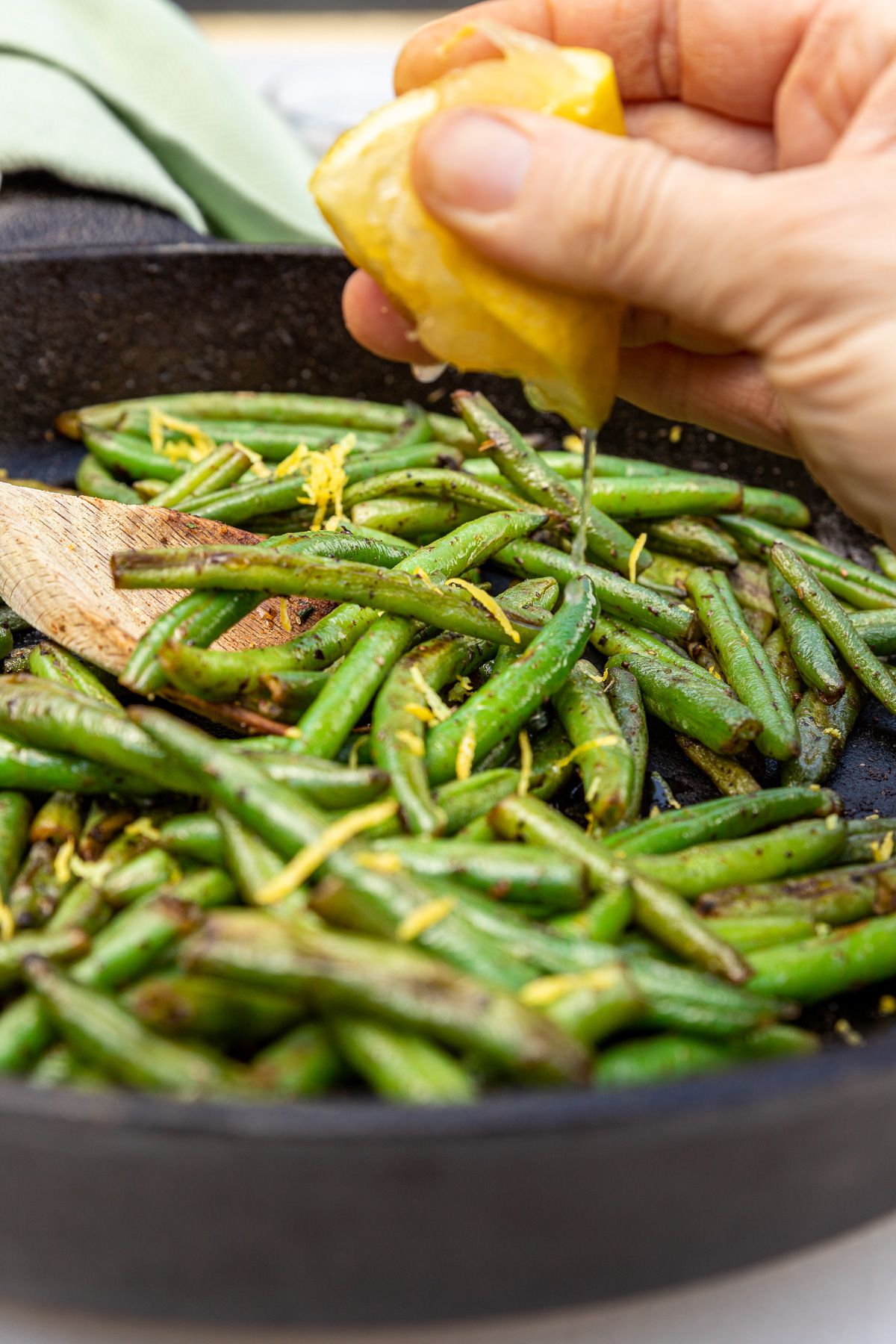 Closeup of someone squeezing half a lemon into a black cast iron pan of sauteed frozen green beans with a pale green tea towel in the background.