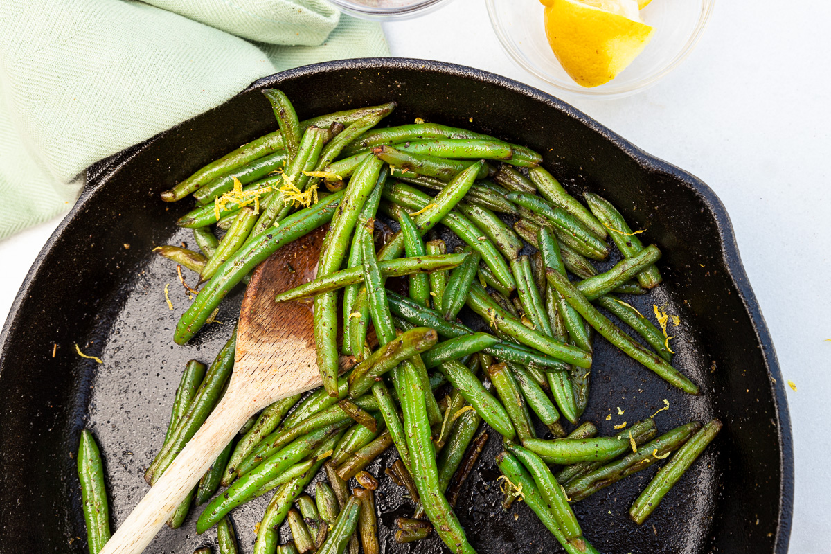 Closeup of overhead view of top half of a black cast iron pan with a wooden spoon in a pan of just-sauteed green beans with a scattering of lemon zest on top and a green tea towel above.