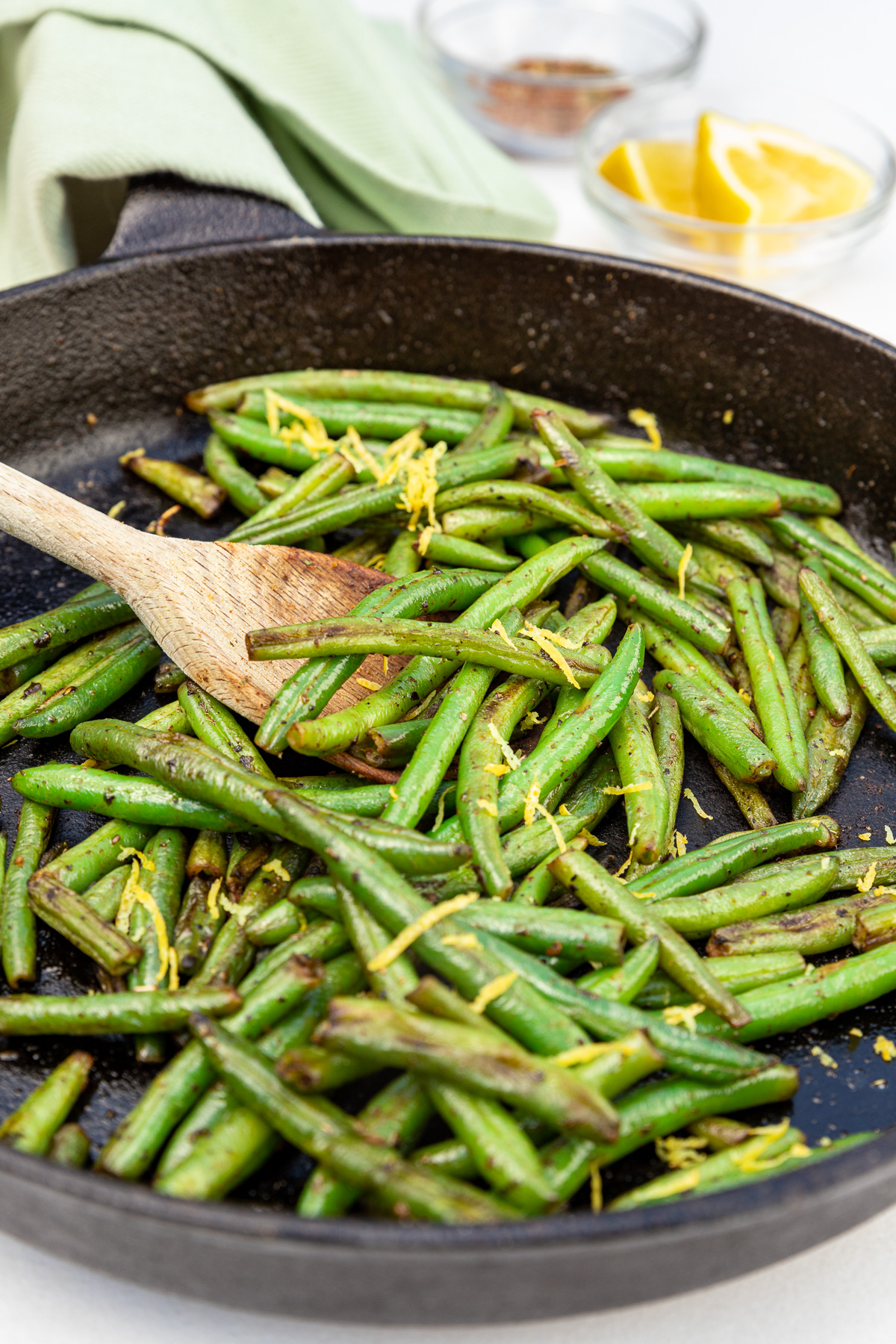 A wooden spoon in a black cast iron pan of just sauteed green beans with lemon zest sprinkled on the top and a green towel and lemon in a bowl in the background.