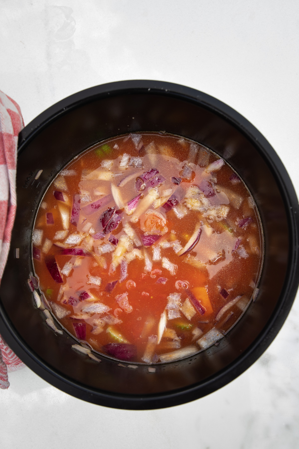 Overhead of all the ingredients for tomato soup in an instant pot on a marble background and with a red and white tea towel.