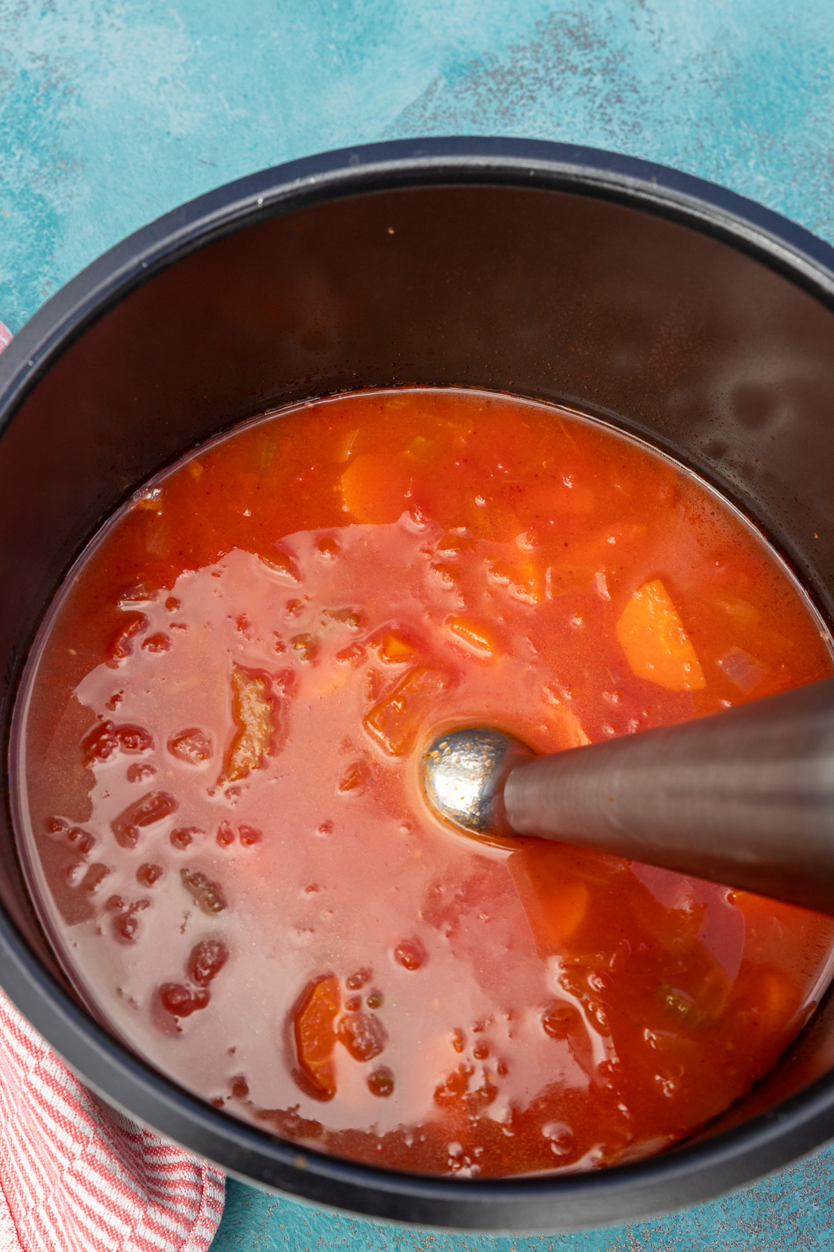 Closeup of immersion blender in tomato soup just about to blend on a blue background with a red and white tea towel.