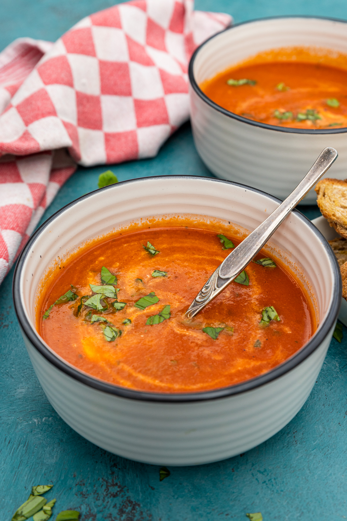 A bowl of instant pot tomato soup with a spoon in and fresh basil on top with another bowl in the background and on a blue table.