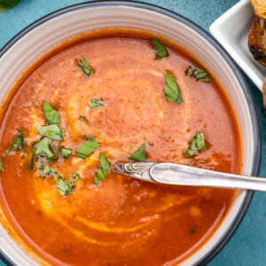Closeup overhead shot of a bowl of instant pot tomato soup with a spoon in and fresh basil on top and with fresh basil in the top left corner.