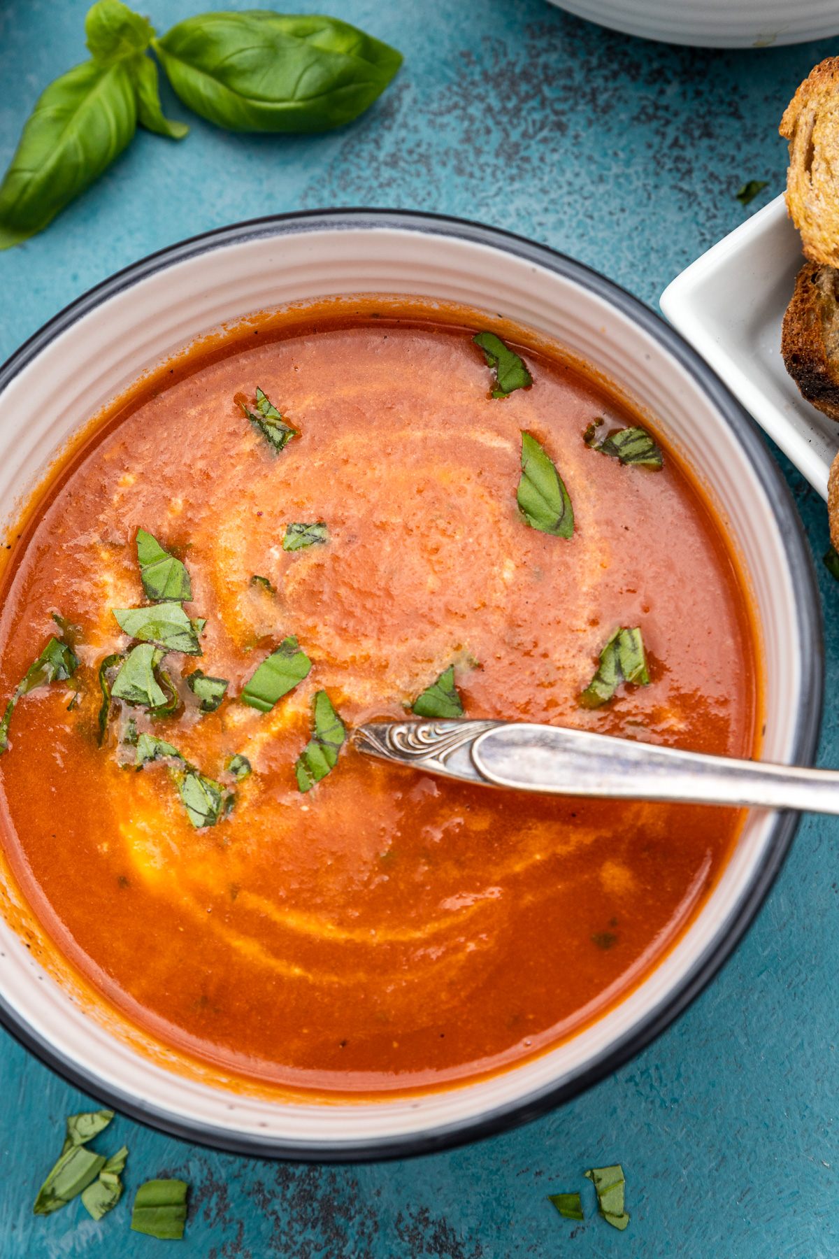 Closeup overhead shot of a bowl of instant pot tomato soup with a spoon in and fresh basil on top and with fresh basil in the top left corner.