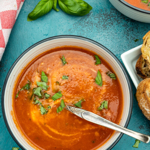 Overhead of a bowl of instant pot tomato soup with a spoon in it and fresh basil on top with more soup in the corner and all on a blue background.
