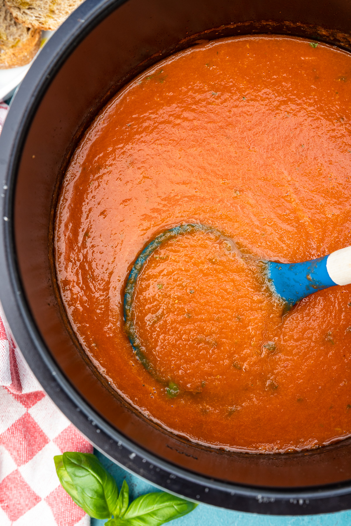 Overhead closeup shot of tomato soup in an instant pot with a blue ladle in it and with a red and white checked tea towel and fresh basil in the corner.